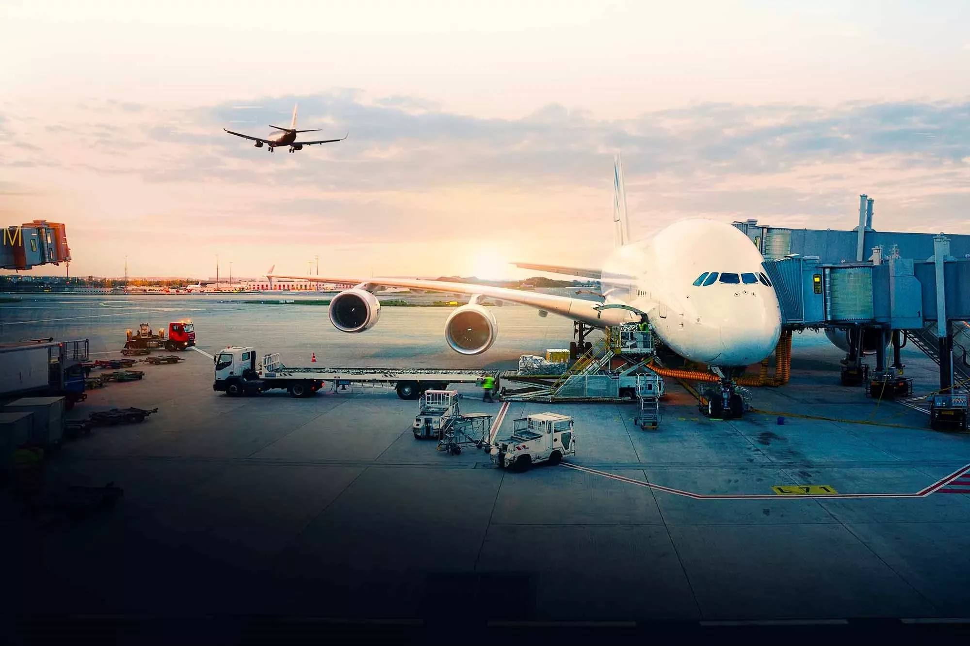 The airport at sunset. A passenger plane is parked on the ground and another one is coming in for landing. Ground Support Equipment is driving on the ground.