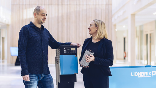 A man and a woman is talking, standing on each side of a blue industrial battery charger.